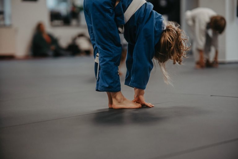 Über uns 6 Kind im blauen Gi führt eine Dehnübung auf der Matte während des Kindertrainings bei Harrycane Luta Livre aus.