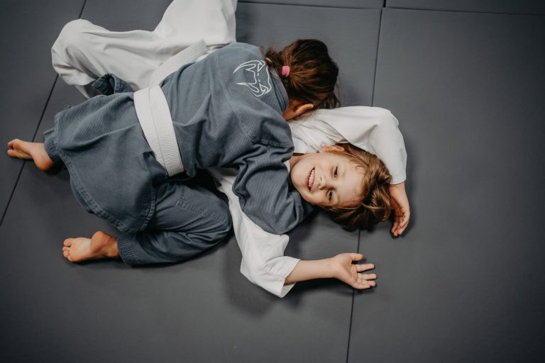 Über uns 19 Zwei Kinder in Luta Livre Trainingsanzügen beim Techniktraining auf der Matte, lächelnd und spielerisch.